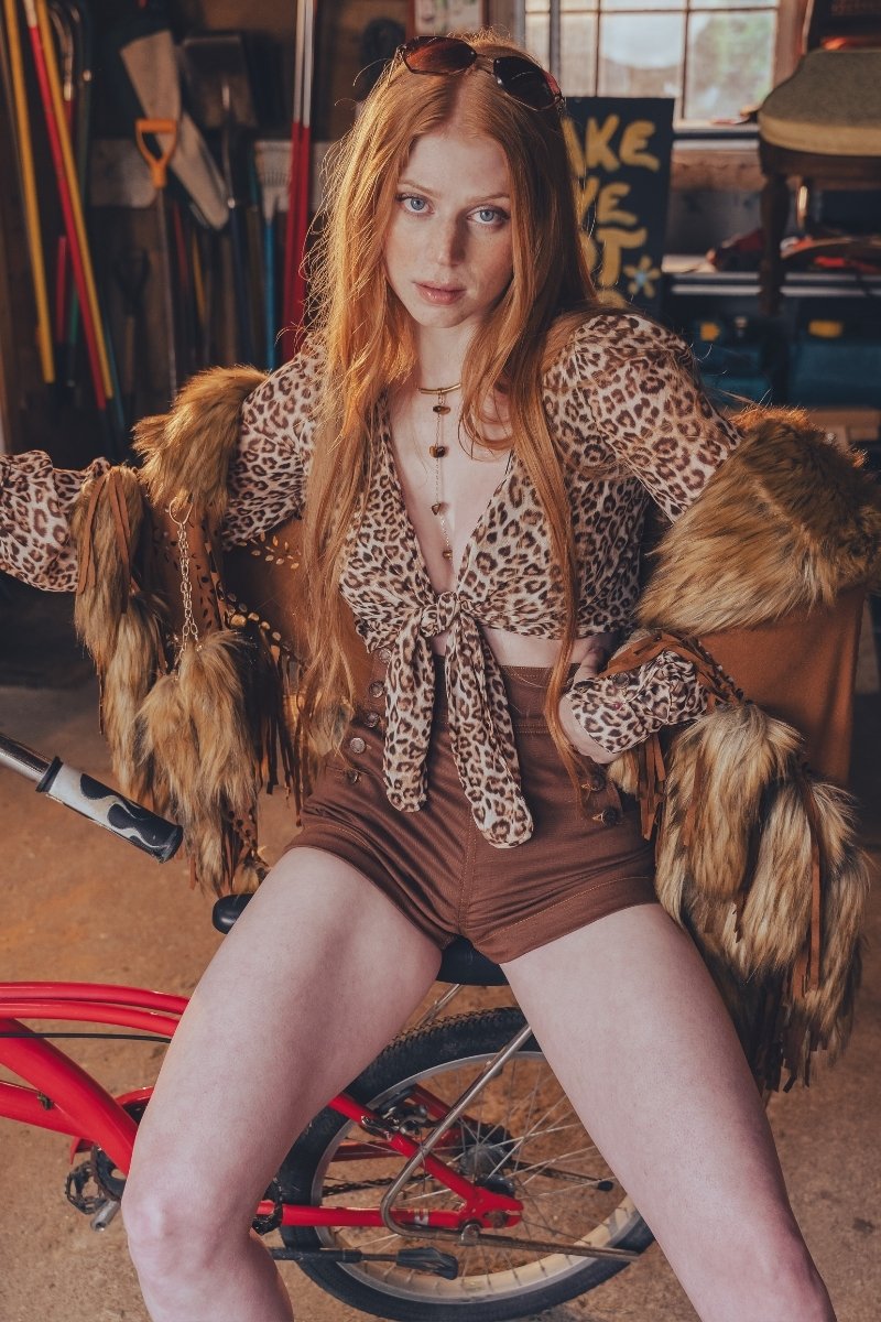 Person wearing a leopard print outfit with fur accents sitting on a red bicycle in a workshop.