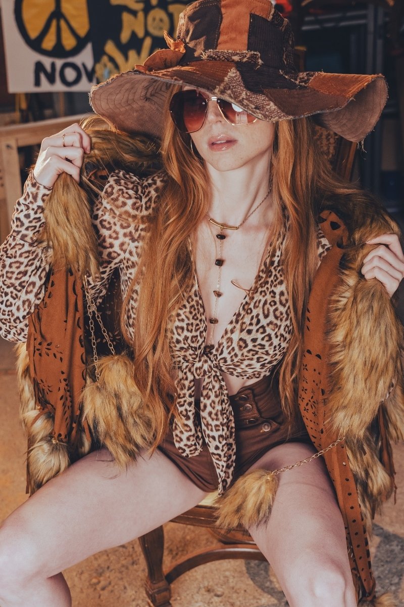 Person in a leopard print outfit with a large hat and sunglasses, sitting on a bench.