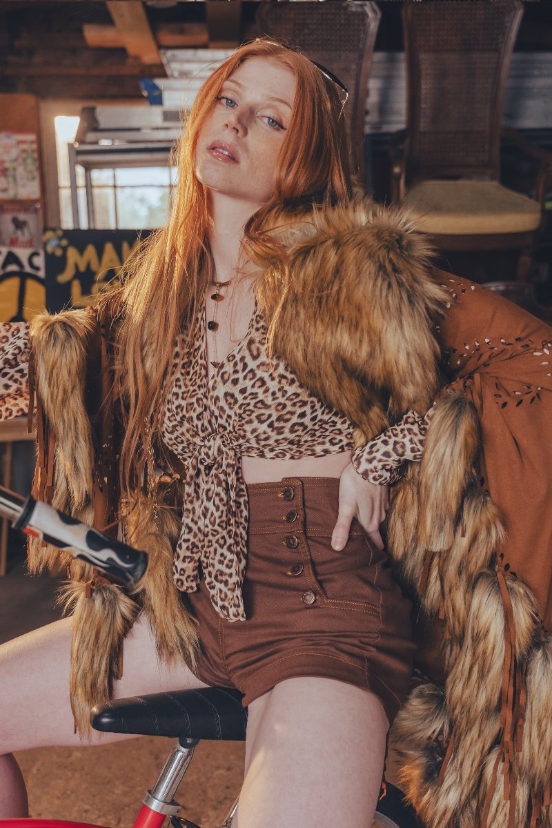 Woman in a fur coat and leopard print top sitting on a chair with a rustic background.