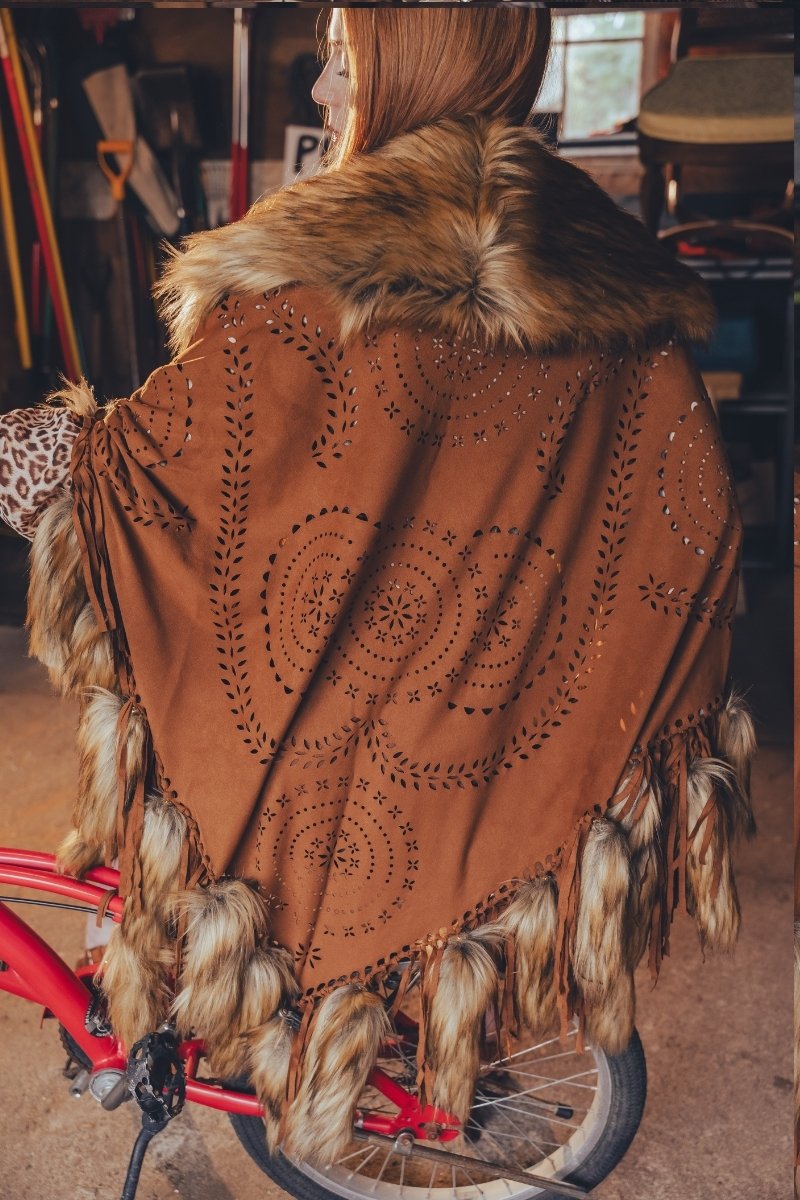Person wearing a brown poncho with fur trim and intricate patterns, standing next to a red bicycle.
