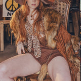 Woman in a vintage-style outfit with a fur stole sitting on a chair in a rustic setting.