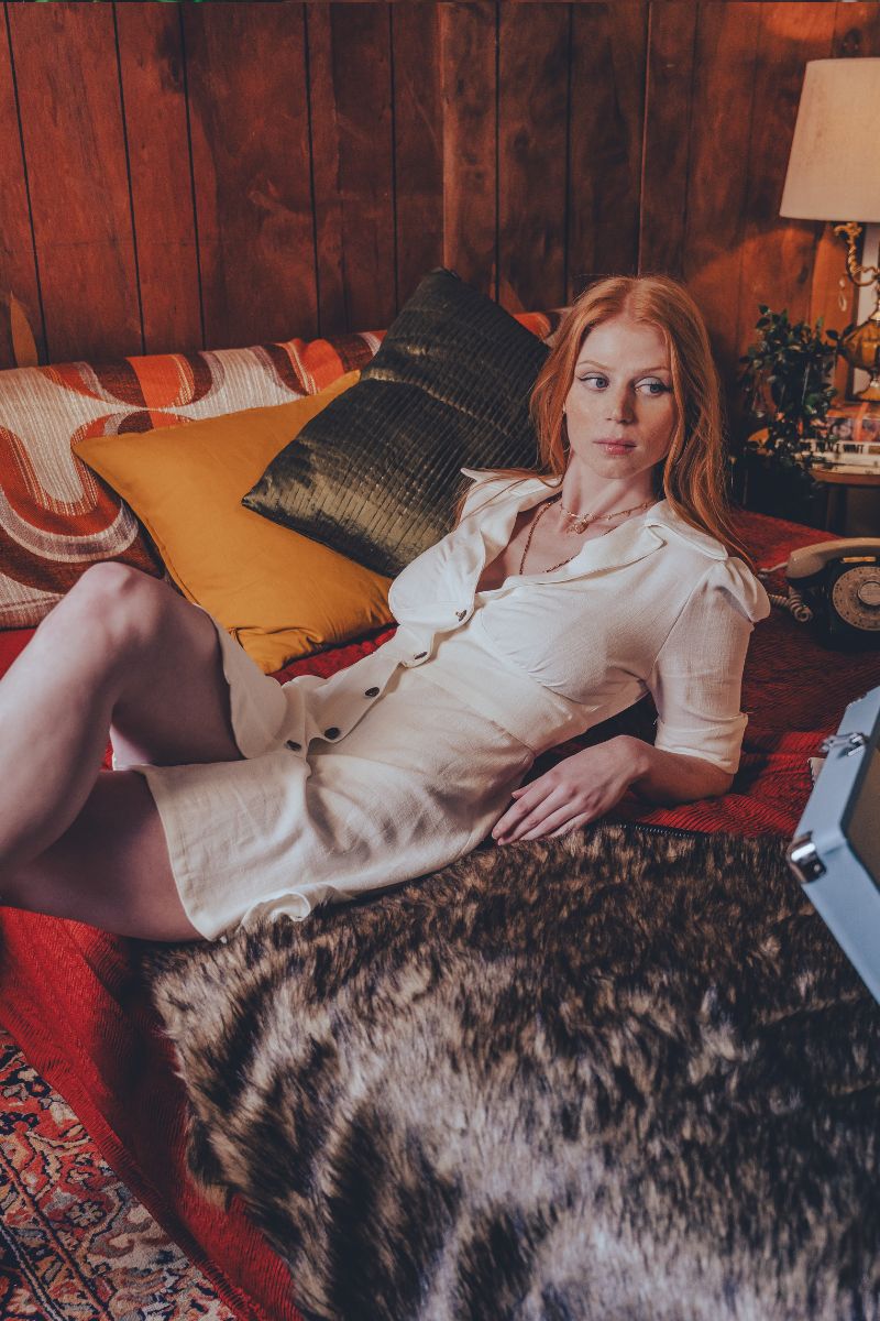 Woman in a white dress lying on a red couch with wooden walls and a lamp in the background