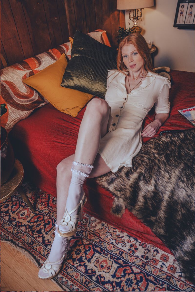 Woman in a white dress and knee-high boots sitting on a red couch with a cat.