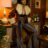A person wearing a brown furry jacket and bell bottoms with a multi-striped pattern, sitting in a room with a wooden background and a plant.