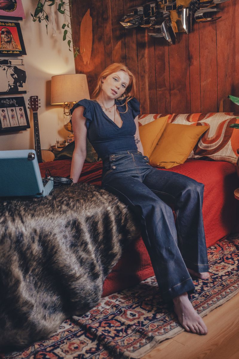 Woman sitting on a couch in a cozy living room with a guitar and lamp in the background.