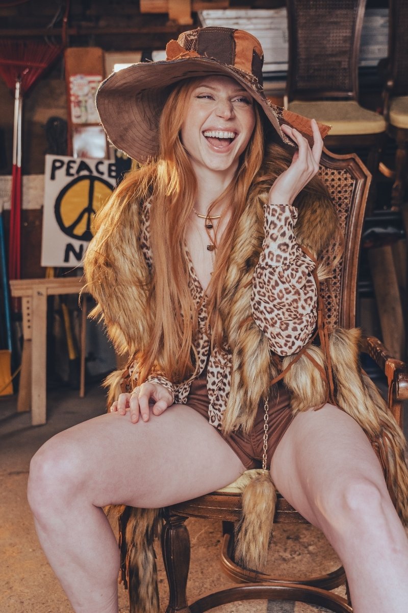 Woman with long red hair wearing a wide-brimmed hat and leopard print top, sitting on a wooden chair.