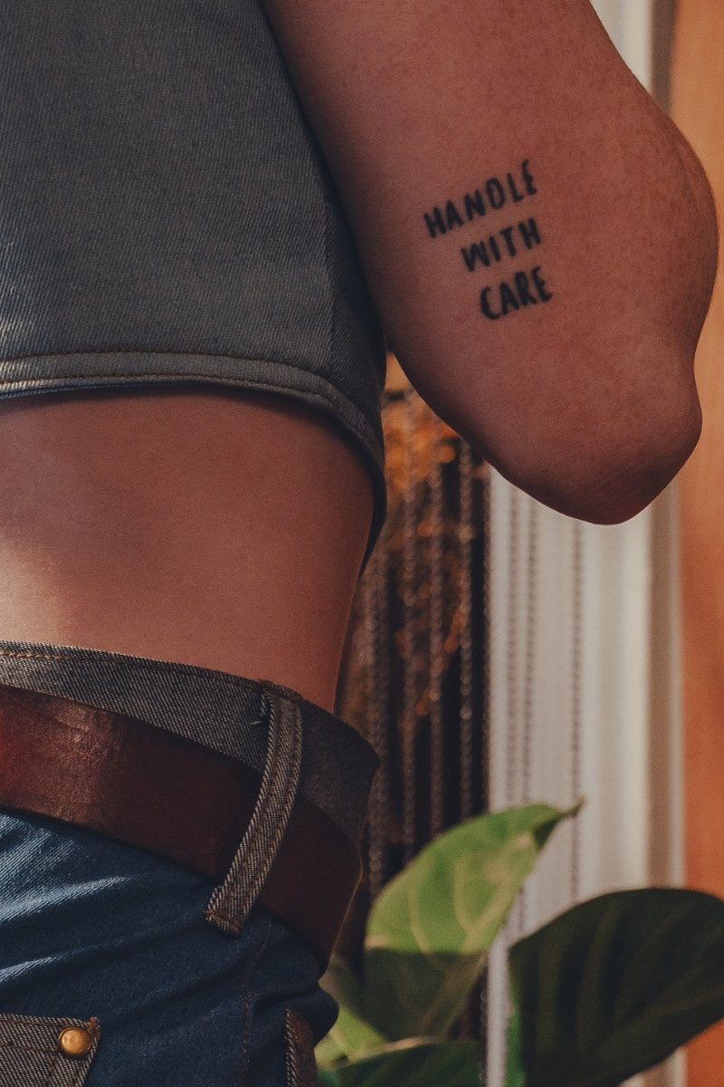 Person wearing a tattoo on their arm with the text 'HANDLE WITH CARE' against a blurred background.