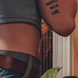 Person wearing a tattoo on their arm with the text 'HANDLE WITH CARE' against a blurred background.