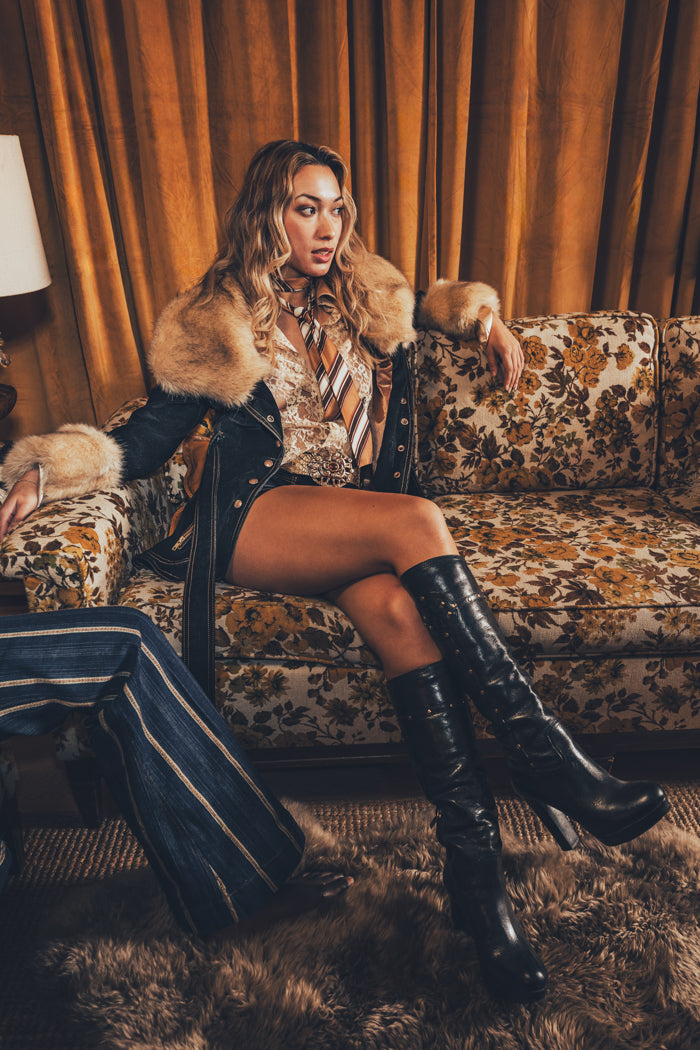 A person wearing a faux fur lined coat with gold satin detailing and bold topstitching, paired with a patterned shirt and shorts.