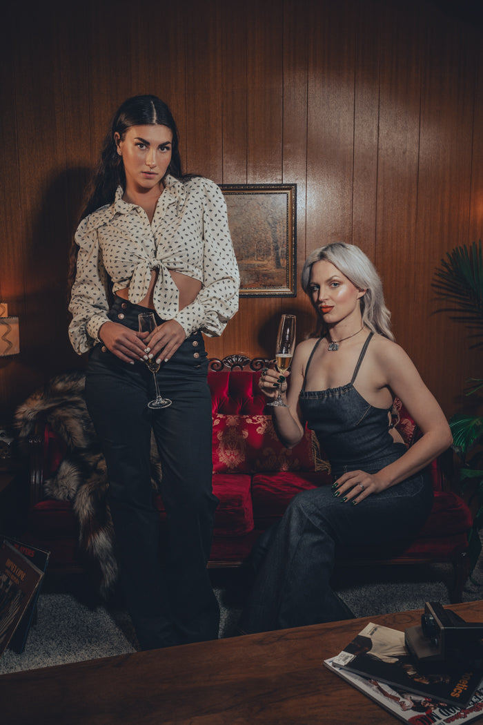 models channels 1970s fashion with flared black trousers and a vest and the other model wears a strappy backless black denim jumpsuit in a groovy home with wood paneling and funky decor.