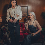 models channels 1970s fashion with flared black trousers and a vest and the other model wears a strappy backless black denim jumpsuit in a groovy home with wood paneling and funky decor.