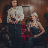 models channels 1970s fashion with flared black trousers and a vest and the other model wears a strappy backless black denim jumpsuit in a groovy home with wood paneling and funky decor.