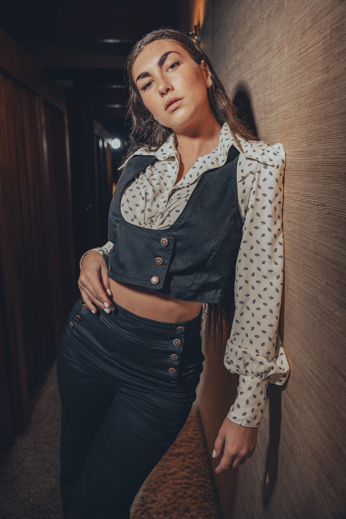 model wearing 70's inspired black bell bottoms and matching vest poses by a record player in a vintage style room