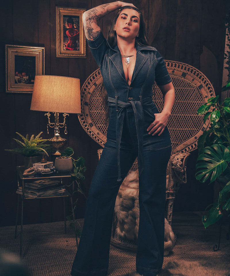 Model wearing a 70s inspired stretch denim jumpsuit with flared legs front zip closure and a plunging neckline styled for a bold retro look