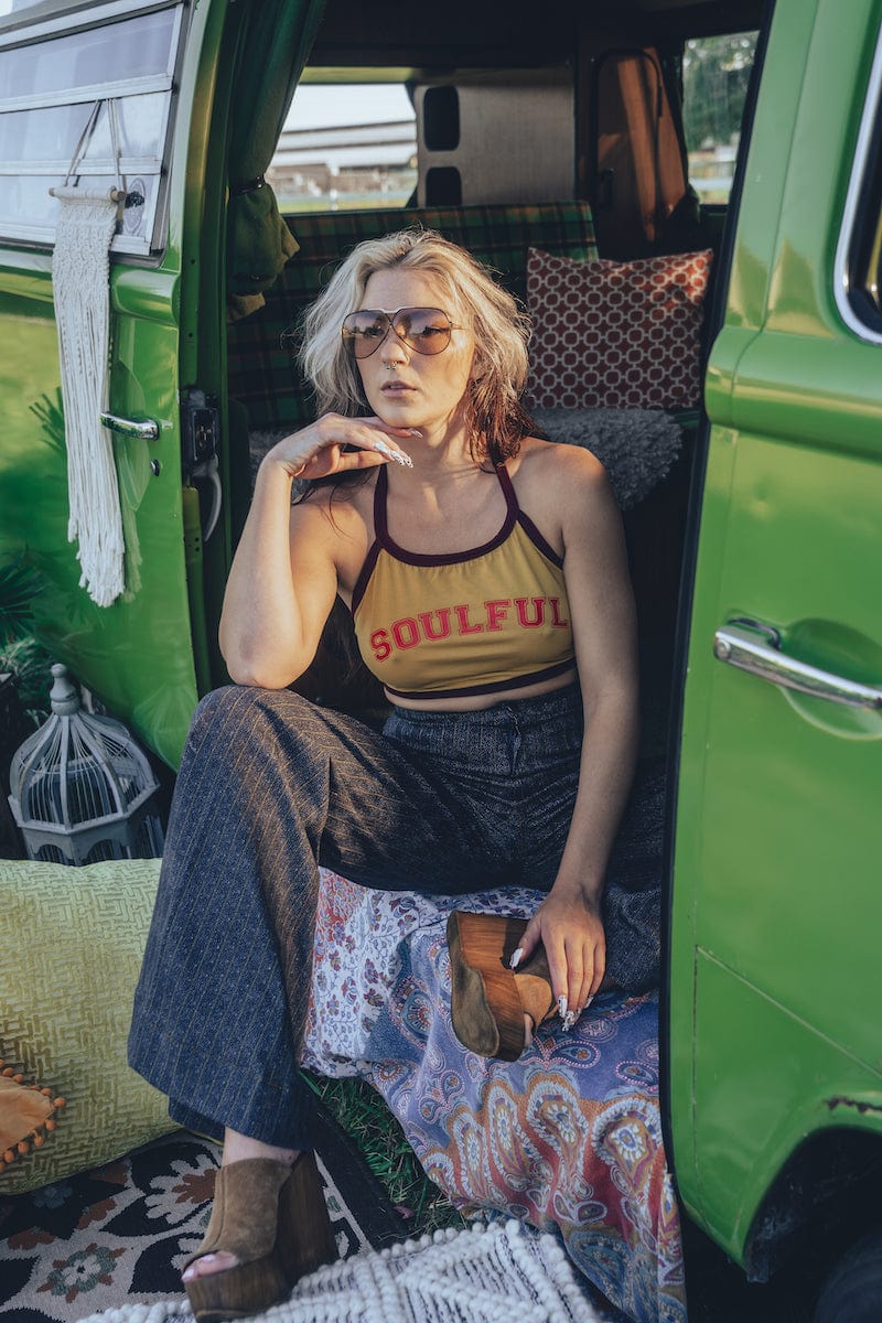 A person wearing a yellow halter top with the word 'SOUULFUL' printed on it, sitting in a green vehicle with their legs dangling outside.