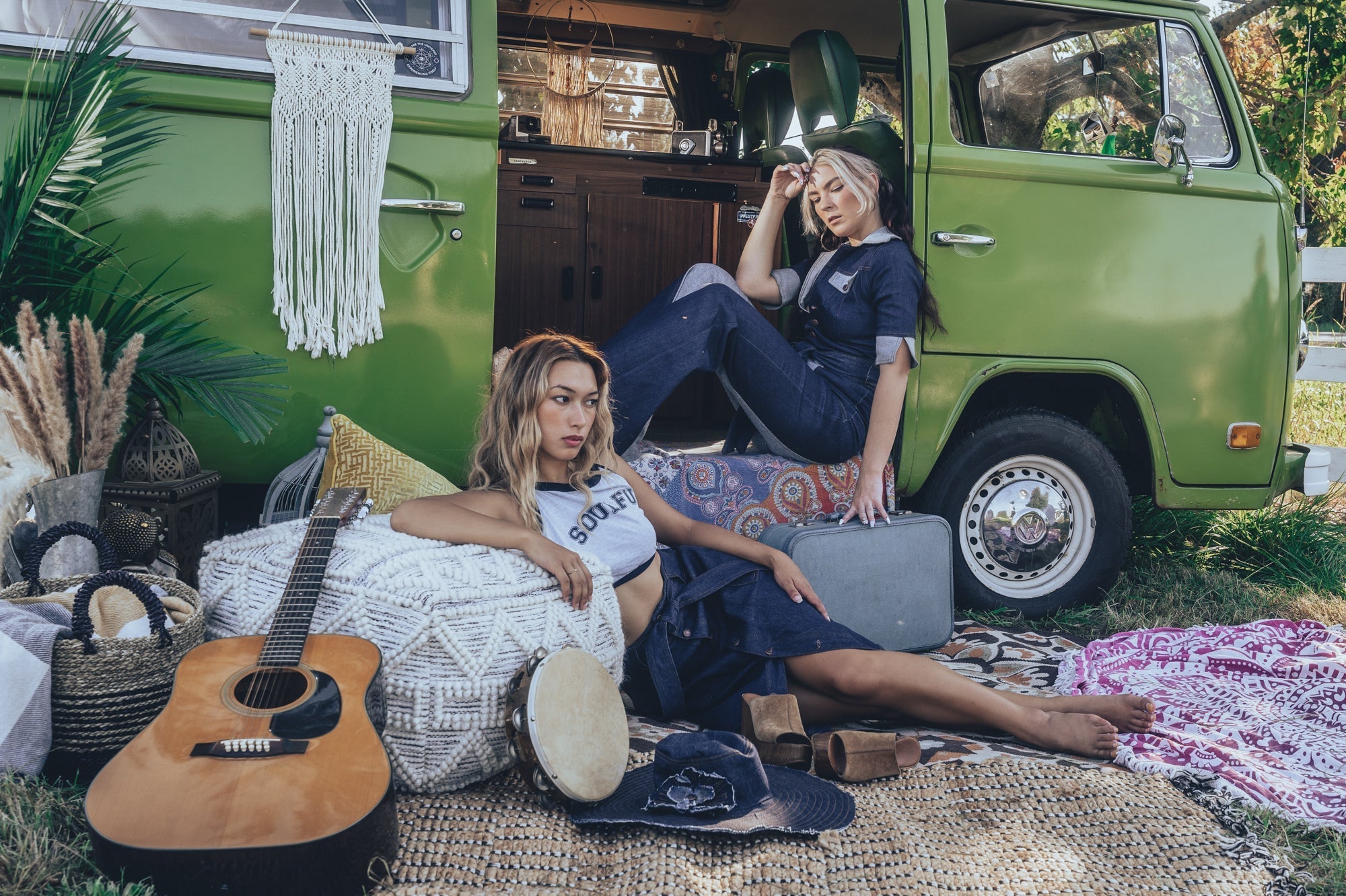 Two girls lounging outside and 1970s VW van with musical instruments one where is a bell bottom jumpsuit the other a BoHo cropped halter top with a denim skirt capturing retro 70s vibes