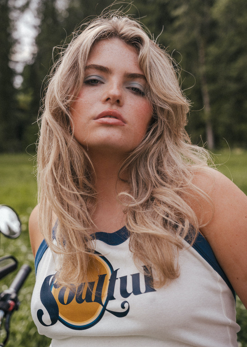 Girl in a 1970s roller skating rink wearing retro sunglasses and a yellow queen halter top with red lettering serving vintage vibes and cool confidence