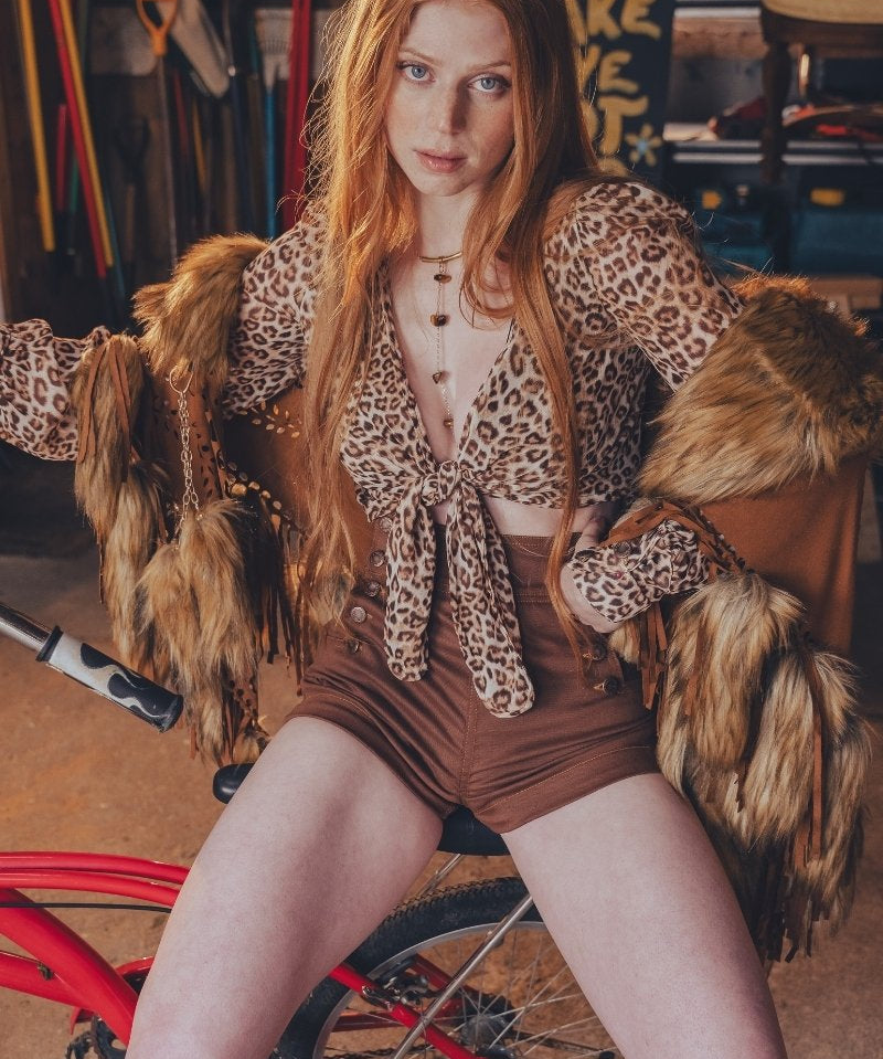 Person wearing a leopard print outfit with fur accents sitting on a red bicycle in a workshop.