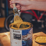 Hand holding a paintbrush over a can of yellow paint on a wooden surface.