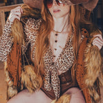 Person in a leopard print outfit with a large hat and sunglasses, sitting on a bench.
