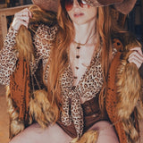 Person in a leopard print outfit with a large hat and sunglasses, sitting on a bench.