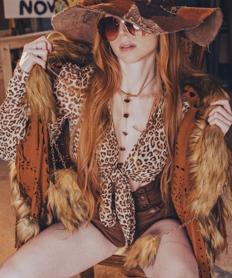 Person in a leopard print outfit with a large hat and sunglasses, sitting on a bench.