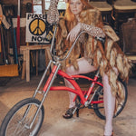 Person sitting on a red bicycle in a cluttered room with chairs and a 'Peace Now' poster.