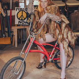 Person sitting on a red bicycle in a cluttered room with chairs and a 'Peace Now' poster.