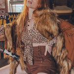 Woman in a fur coat and leopard print top sitting on a chair with a rustic background.