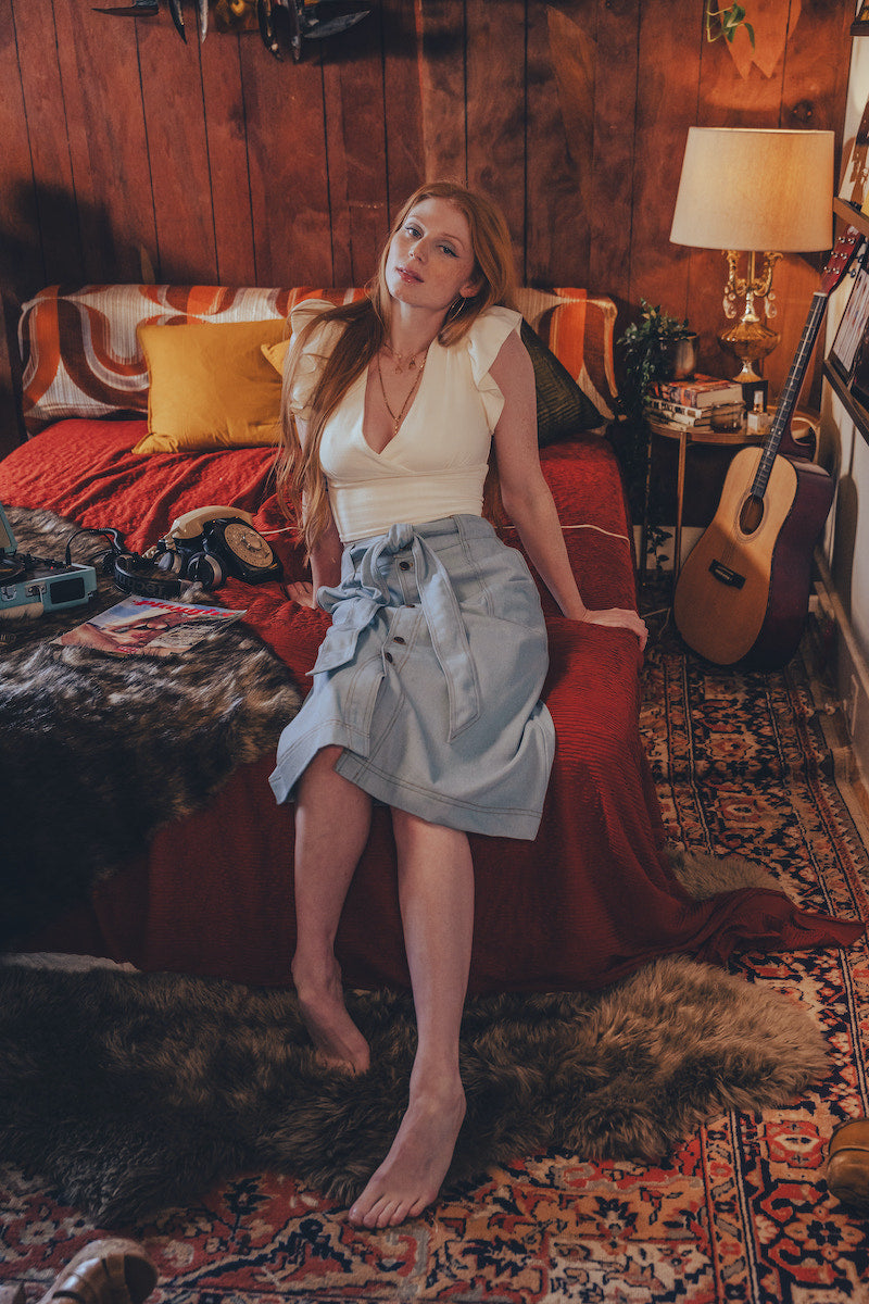 Woman sitting on a bed in a cozy room with wooden walls and a guitar.