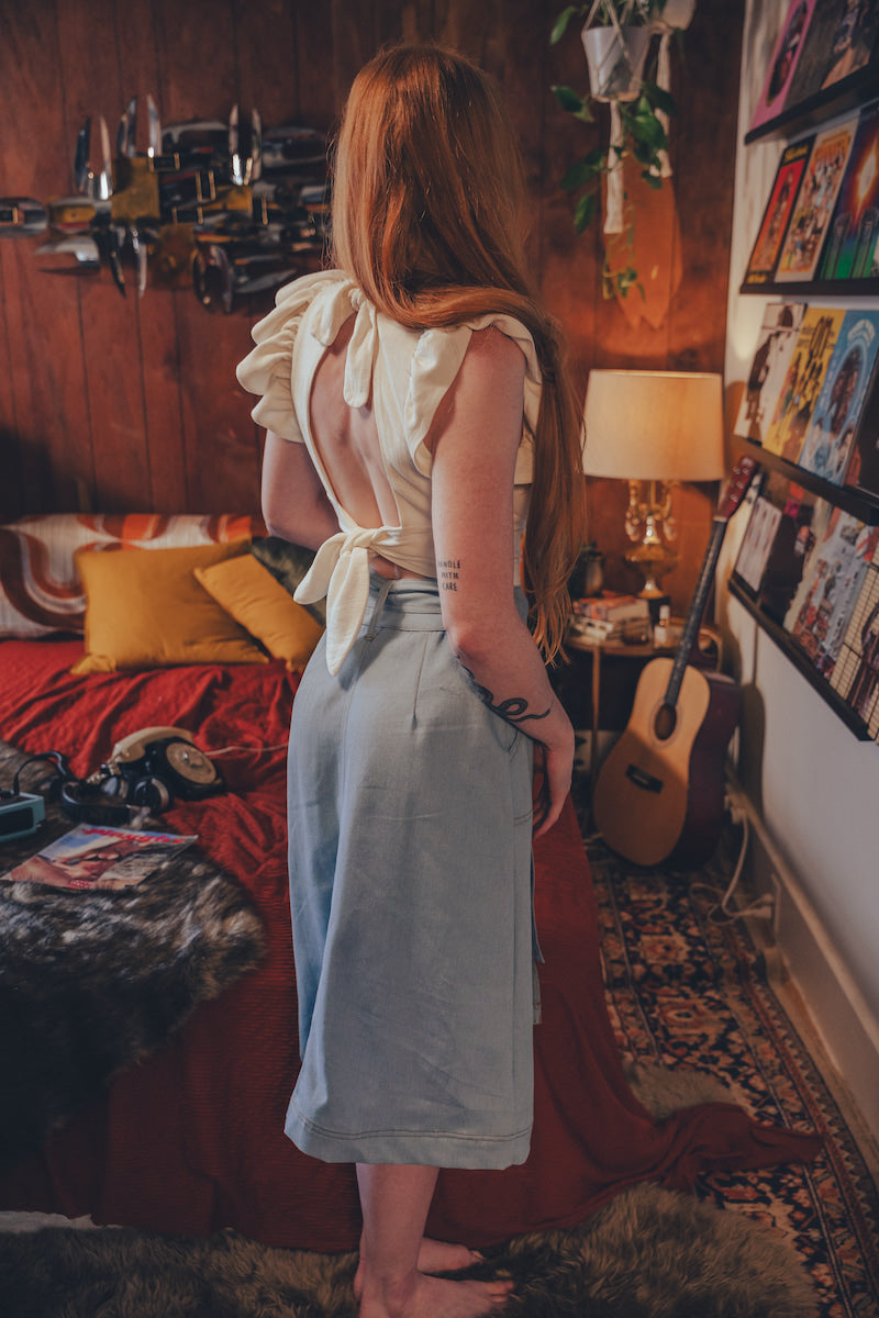 Woman in a light blue dress standing in a cozy bedroom with a guitar and books.