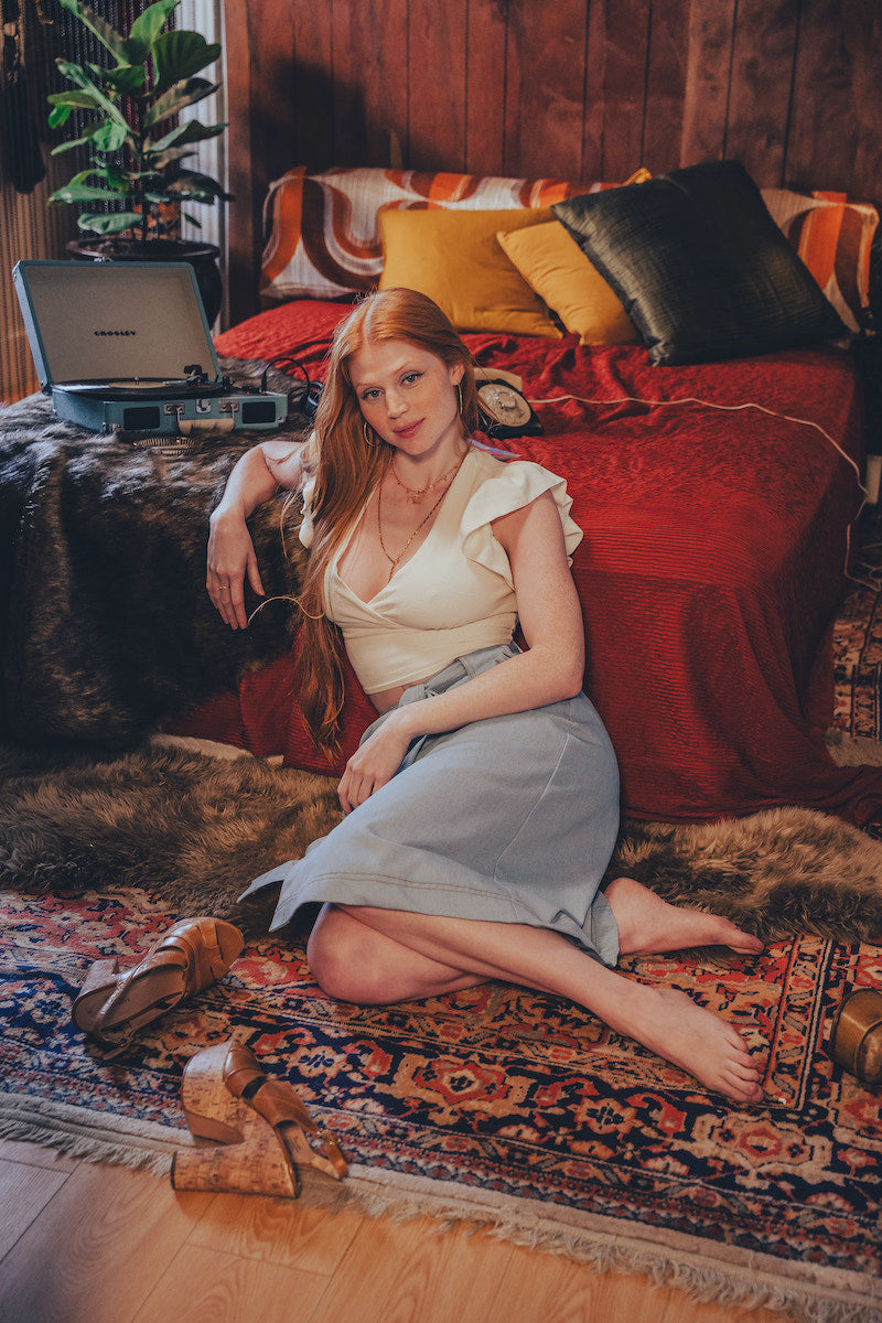 Woman sitting on a patterned rug in a cozy room with a bed and plants.