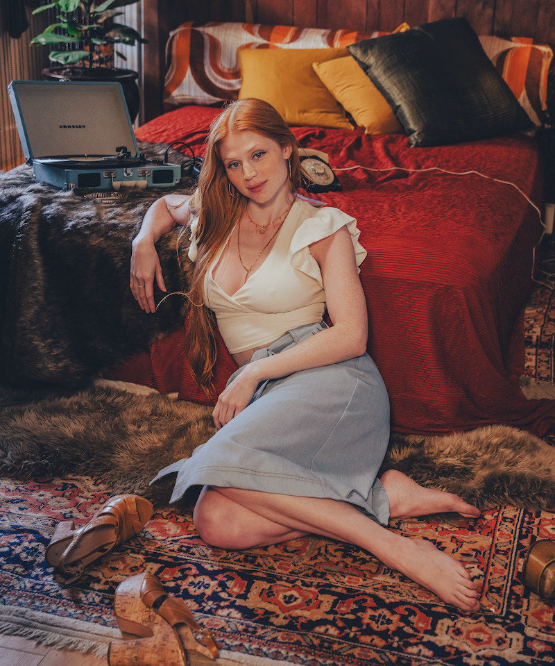 Woman sitting on a patterned rug in a cozy room with a bed and plants.