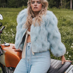 Woman in light blue fur coat and jeans sitting on a motorcycle in a forest setting wearing Soul Sister since 1969