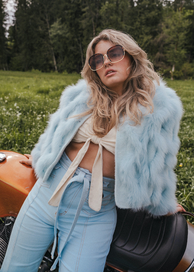 Woman in light blue fur coat and jeans sitting on a motorcycle in a field wearing Soul Sister since 1969.