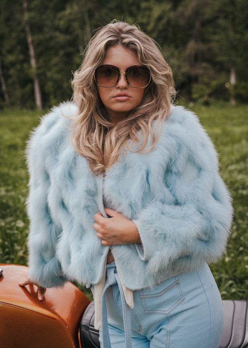 Woman wearing a light blue fur coat and sunglasses outdoors wearing Soul Sister since 1969.