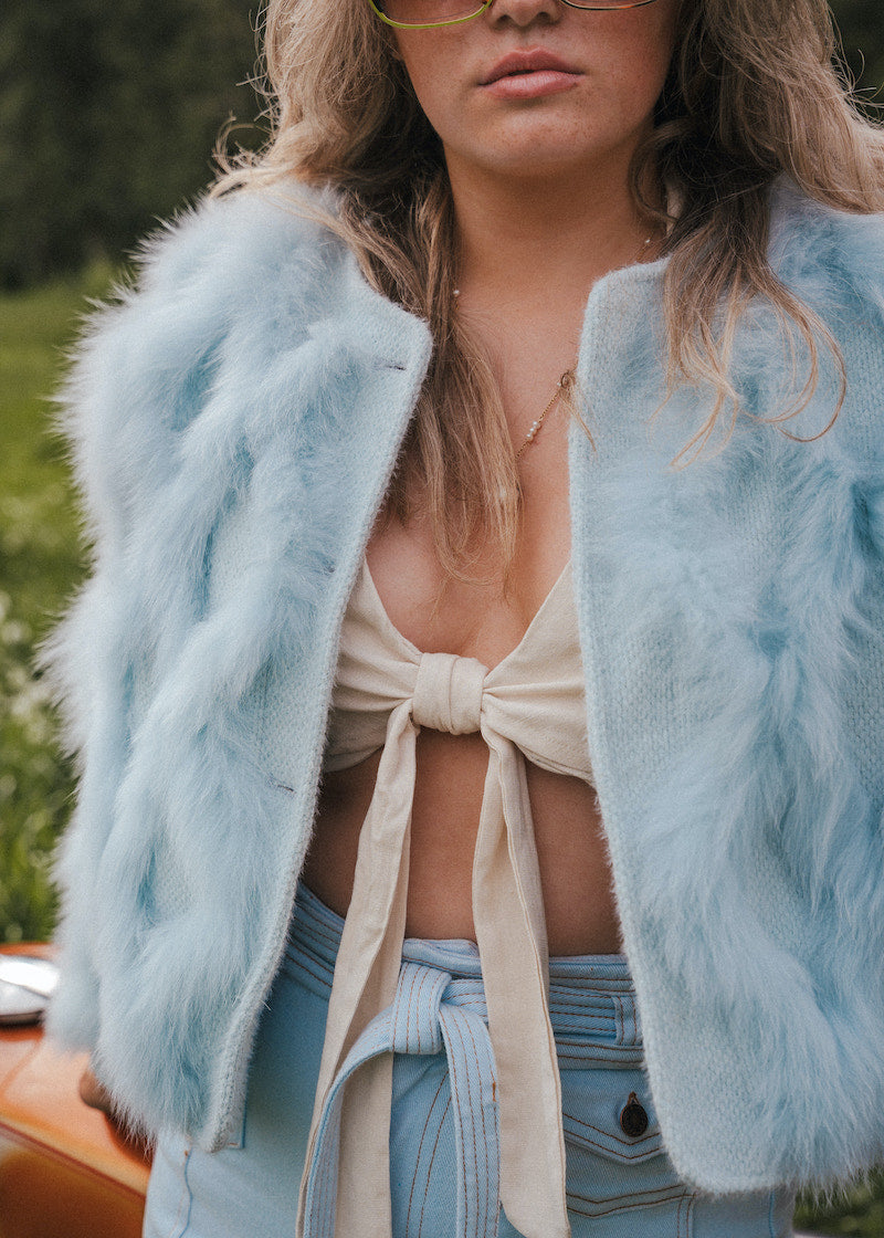 Person wearing a light blue fluffy jacket over a beige top and denim pants, with a blurred natural background wearing Soul Sister since 1969.