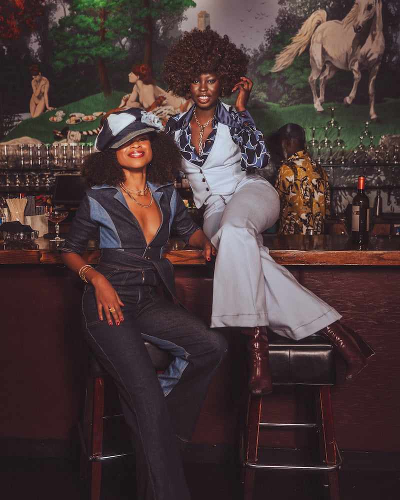 Two people sitting at a bar with a vintage painting of nude figures in the background