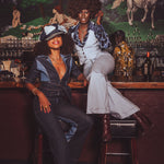 Two people sitting at a bar with a vintage painting of nude figures in the background