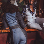 Two women sitting at a bar, one wearing a denim jumpsuit and the other in a patterned outfit.