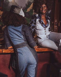 Two women sitting at a bar, one wearing a denim jumpsuit and the other in a patterned outfit.