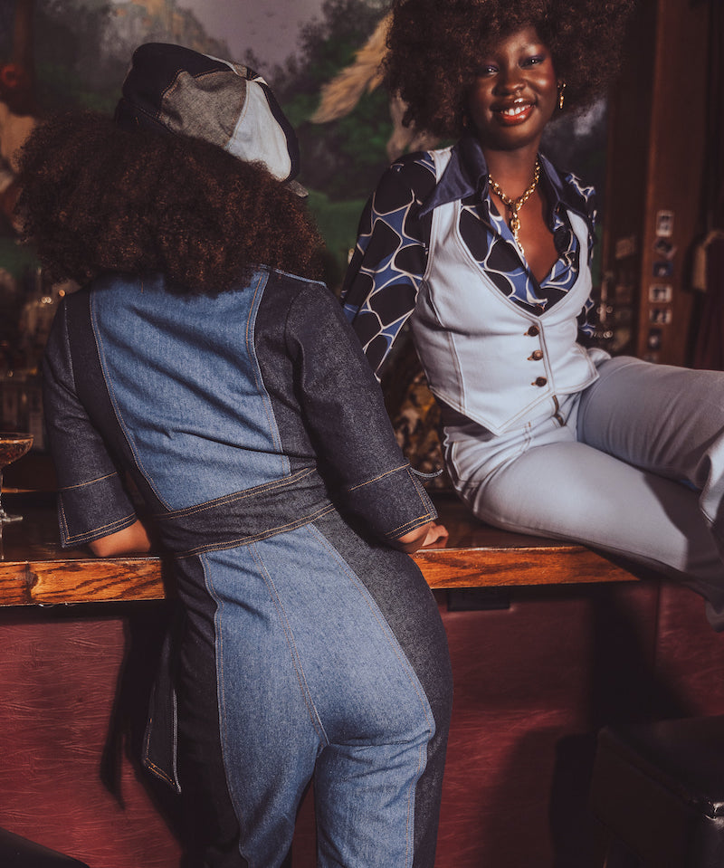 Two women sitting at a bar, one wearing a denim jumpsuit and the other in a patterned outfit.