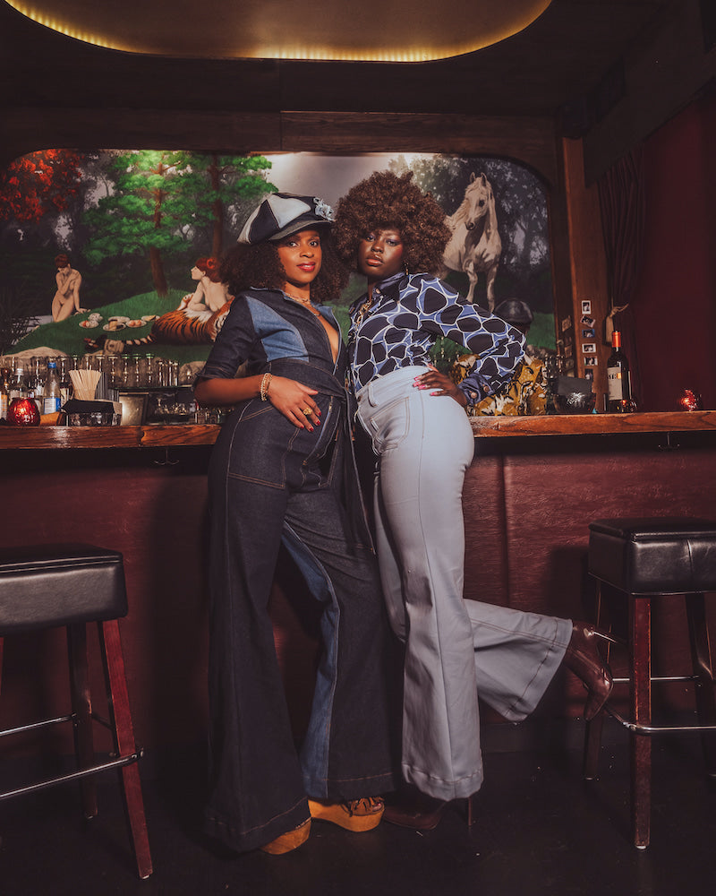 Two women posing in a bar with a painting of a picnic scene on the wall.