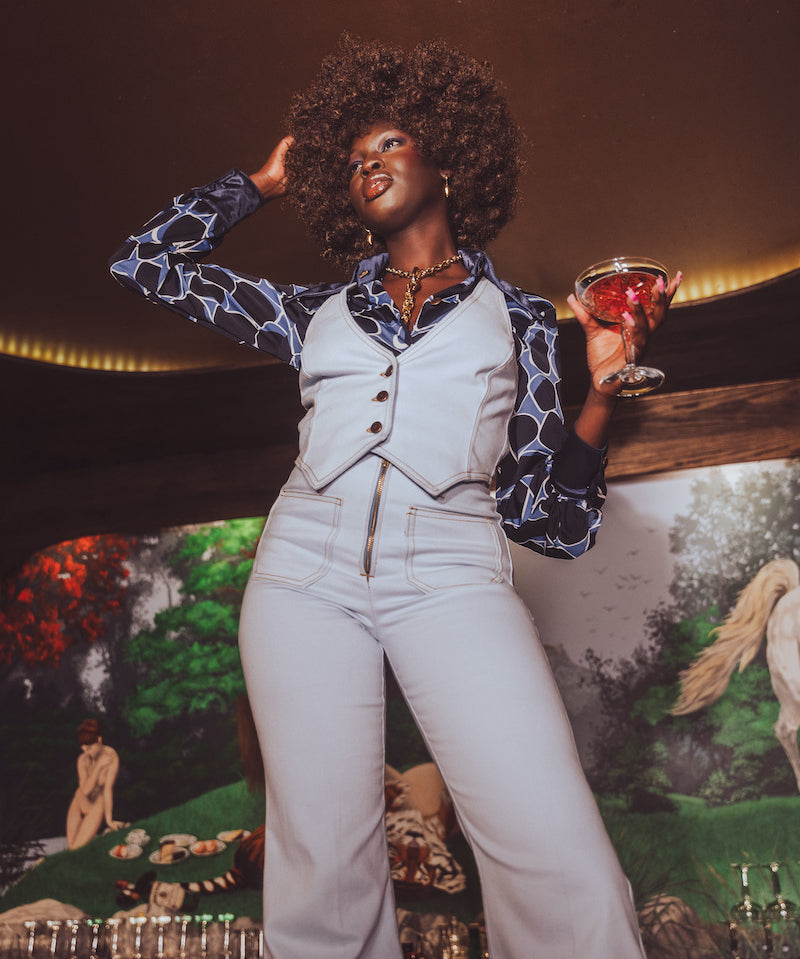 Woman in a stylish outfit holding a drink against a mural background