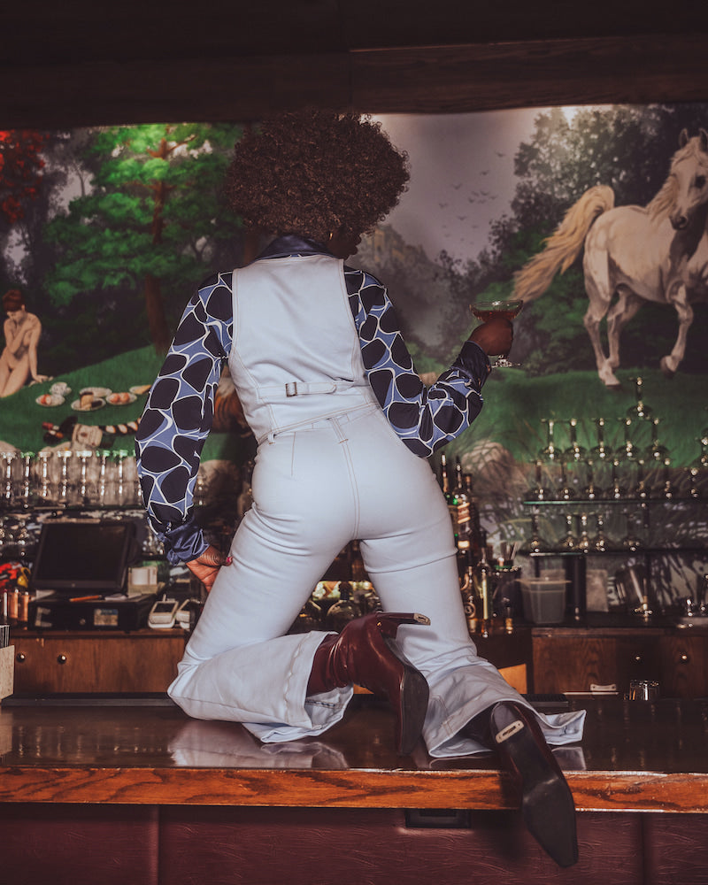 Person in white overalls and patterned shirt leaning against a bar with a horse painting in the background