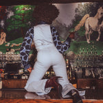 Person in white overalls and patterned shirt leaning against a bar with a horse painting in the background