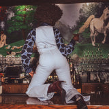 Person in white overalls and patterned shirt leaning against a bar with a horse painting in the background