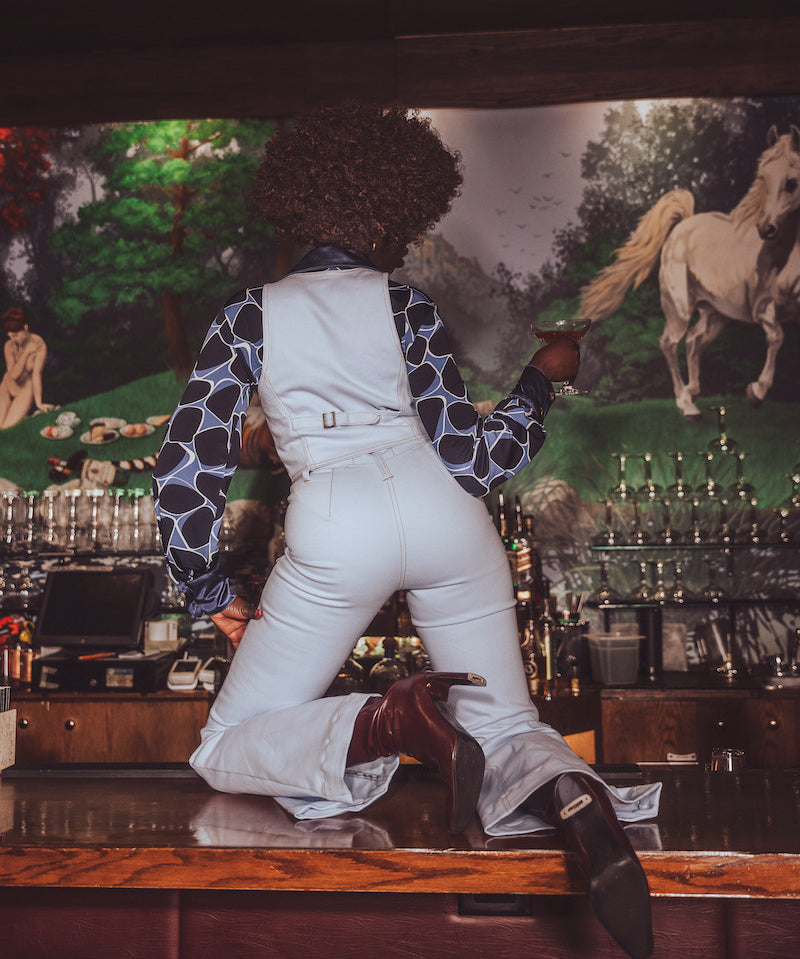 Person in white overalls and patterned shirt leaning against a bar with a horse painting in the background