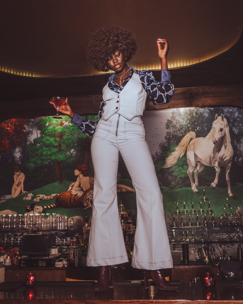 Person in a pale blue retro 70s outfit standing in front of a decorative wall with horse paintings.