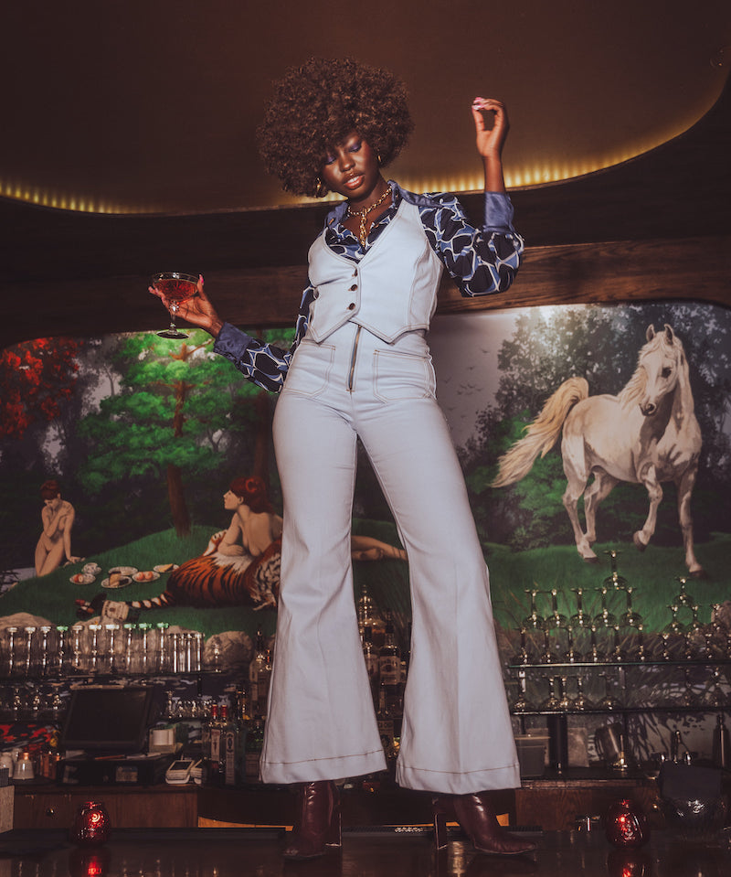 Person in a pale blue retro 70s outfit standing in front of a decorative wall with horse paintings.